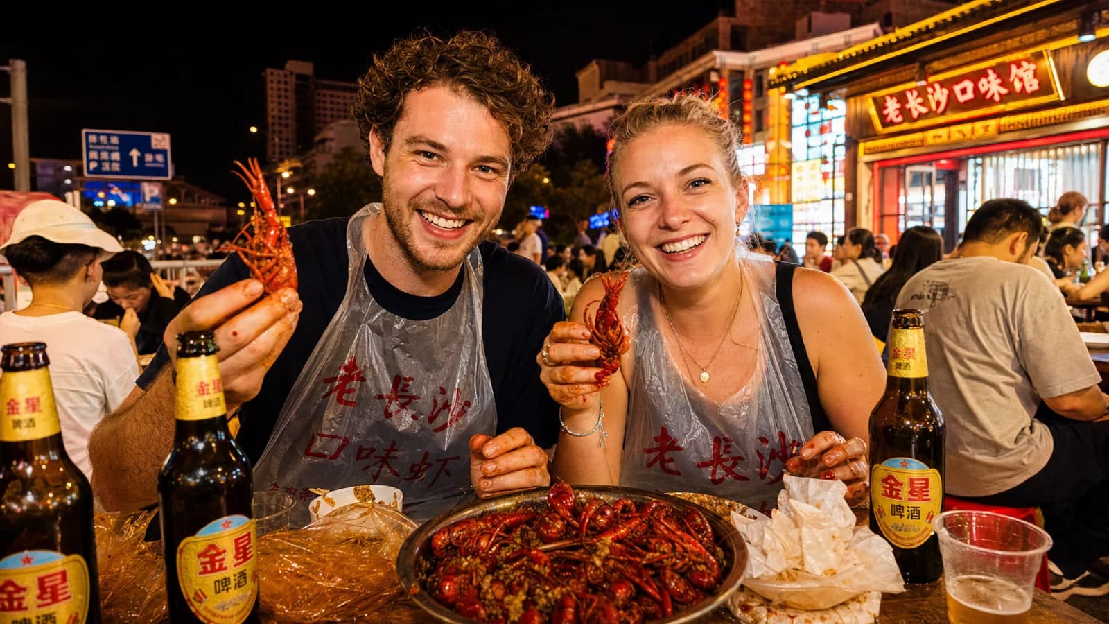 Travelers in plastic bibs sharing a messy platter of spicy Changsha crayfish at a night-market table